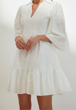 Vrouw met een witte jurk met V-hals, ballonmouwen, gebloemd ajour kant en een gerimpelde volantzoom, staand met de handen gevouwen.