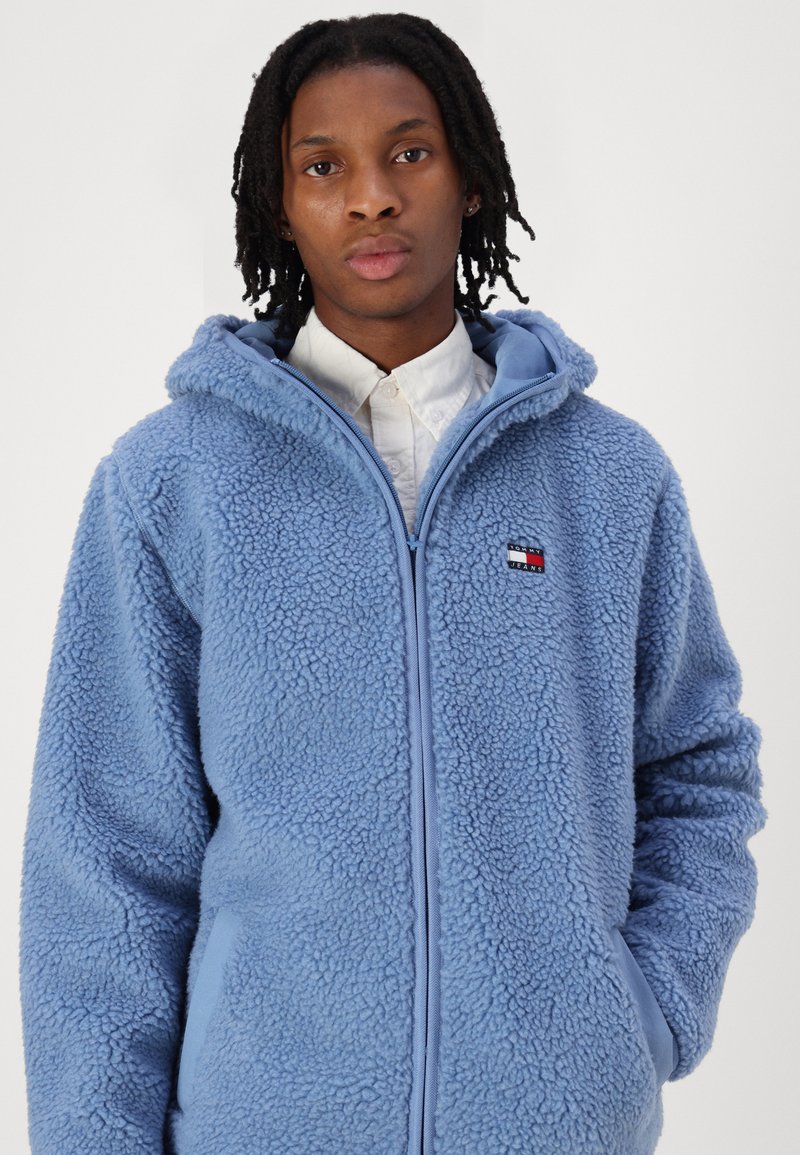 Himmelblauer Fleece-Hoodie mit strukturiertem Außenmaterial, vollständigem Reißverschluss und Fronttaschen. Verfügt über ein Logopatch auf der linken Brust.