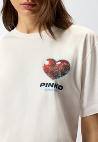 Bjela pamučna majica s velikim crvenim grafičkim oblikom srca koji ima sjajnu teksturu, a ispod toga je otisnuto "PINKO SURREAL LOVE" u crnoj i plavoj boji.