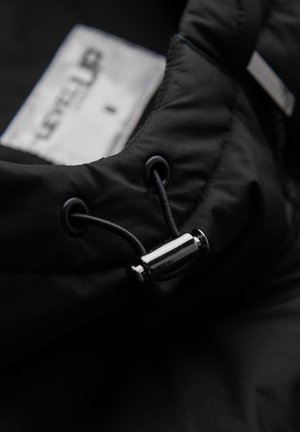 Tessuto della giacca imbottita nera con cordoncino e fermo metallico, presenta un'etichetta con il testo "LEVEL UP" all'interno.