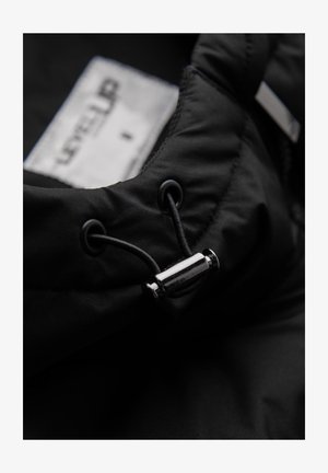 Tissu de veste matelassée noire avec un cordon de serrage et un stoppeur métallique, comportant une étiquette avec le texte "LEVEL UP" à l'intérieur.