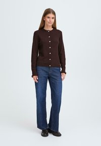 Brauner Cardigan mit Rundhalsausschnitt, langen Ärmeln und Knopfverschluss, kombiniert mit blauen Jeans und schwarzen Schuhen.