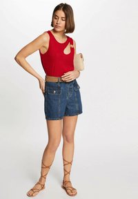 Rotes gestricktes ärmelloses Oberteil mit Ausschnitt, Jeansshorts mit zwei Vordertaschen, hellbra Gürtel und braune Riemchensandalen. Trägt eine beigen Clutch.