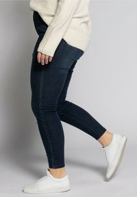 Jeans skinny de denim en azul oscuro, con una cintura alta, bolsillos laterales y un ligero desgastado; combinados con zapatillas blancas.