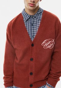 Roter gestrickter Cardigan mit einem tiefen V-Ausschnitt, Knopfleiste und einem weißen grafischen Detail. Getragen über einem blauen karierten Hemd mit Knöpfen.