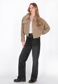 Giacca cropped color tan con due tasche sul petto, abbinata a jeans neri a vita alta. Il modello indossa stivali platform neri. Tessuto liscio e vestibilità casual.