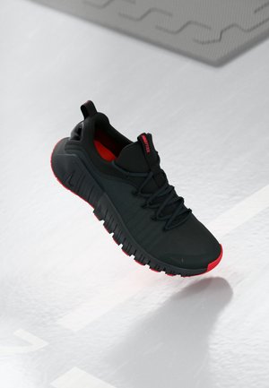 Baskets de sport noires avec semelle rouge et détails, design bas et semelle extérieure segmentée et flexible, présentées flottant au-dessus d'une surface réfléchissante.
