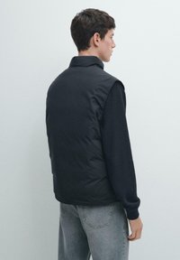 Czarna pikowana kamizelka z wysokim kołnierzem, z gładkim zewnętrznym materiałem i bez rękawów, noszona na teksturowanym ciemnym swetrze.