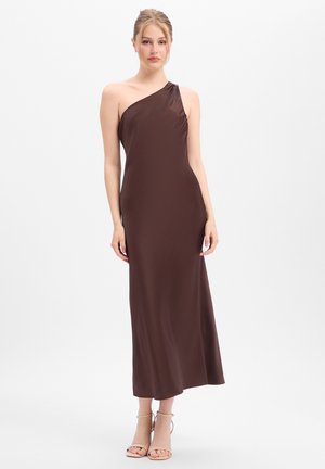 Vrouw draagt een mouwloze, donkerbruine, asymmetrische lange jurk met één schouder en beige open tenen hakken, staand tegen een effen witte achtergrond.