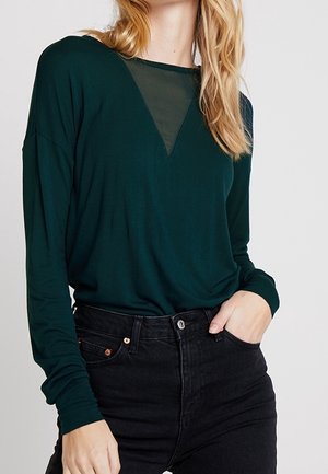 Femme portant un haut à manches longues vert foncé avec une insertion transparente en forme de triangle au niveau de l'encolure et un jean noir taille haute, main dans la poche.
