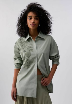 Donna con capelli ricci che indossa una camicia verde chiaro a maniche lunghe con ricamo di foglie e una gonna plissettata, che posa contro uno sfondo semplice.