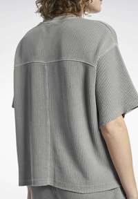 Šedý žebrovaný top s krátkým rukávem, který se vyznačuje volným střihem, kulatým výstřihem a jemnými švy na zádech, které zdůrazňují texturovanou látku.