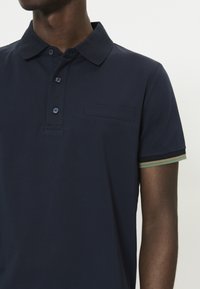 Polo azul marino con cuello clásico, tres botones, un bolsillo delantero y detalles a rayas en verde y beige en las mangas.