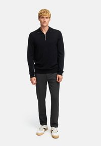 Zwarte trui met ritssluiting, gemaakt van zachte gebreide stof, gecombineerd met grijze broek. Witte sneakers met een beige zool maken de outfit af.