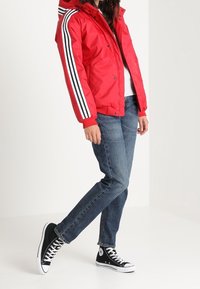 Rød polstret jakke med sorte og hvide stribede ærmer, båret over en grå skjorte og parret med blå jeans og sorte høj-top sneakers.