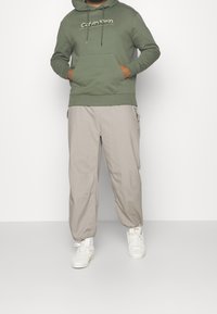 Felpa con cappuccio verde in cotone, dotata di tasca frontale e lacci, abbinata a pantaloni cargo beige e sneakers bianche.