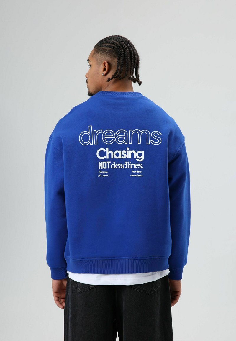 Sweat-shirt bleu avec un grand texte blanc au dos : "dreams Chasing NOT deadlines." Poignets et ourlet côtelés, avec une coupe décontractée.