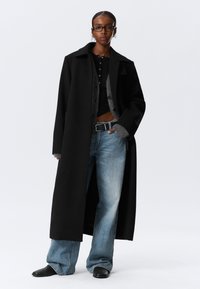 Zwarte lange jas met een gestructureerde kraag, gelaagd over een grijze knoop cardigan. Lichtblauwe flared jeans en zwarte puntige laarzen.
