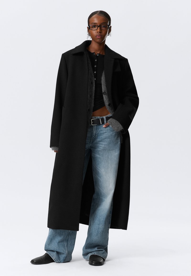 Zwarte lange jas met een gestructureerde kraag, gelaagd over een grijze knoop cardigan. Lichtblauwe flared jeans en zwarte puntige laarzen.