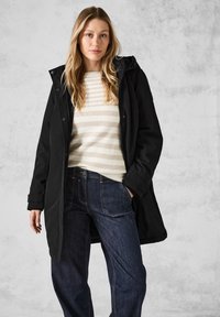 Manteau long noir imperméable avec capuche, sur un pull rayé crème et beige. Une paire de jeans bleu foncé est visible en bas.