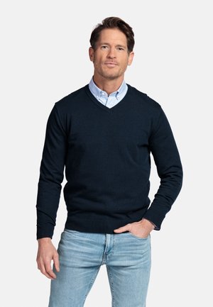 Man draagt een marineblauwe V-hals trui over een lichtblauw overhemd, met lichtblauwe spijkerbroek, staat met één hand in de zak.