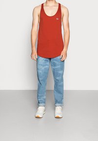 Camiseta sin mangas de algodón rojo con cuello redondo, combinada con jeans de mezclilla azul claros, arremangados en los puños; el modelo lleva zapatillas blancas con detalles en naranja.