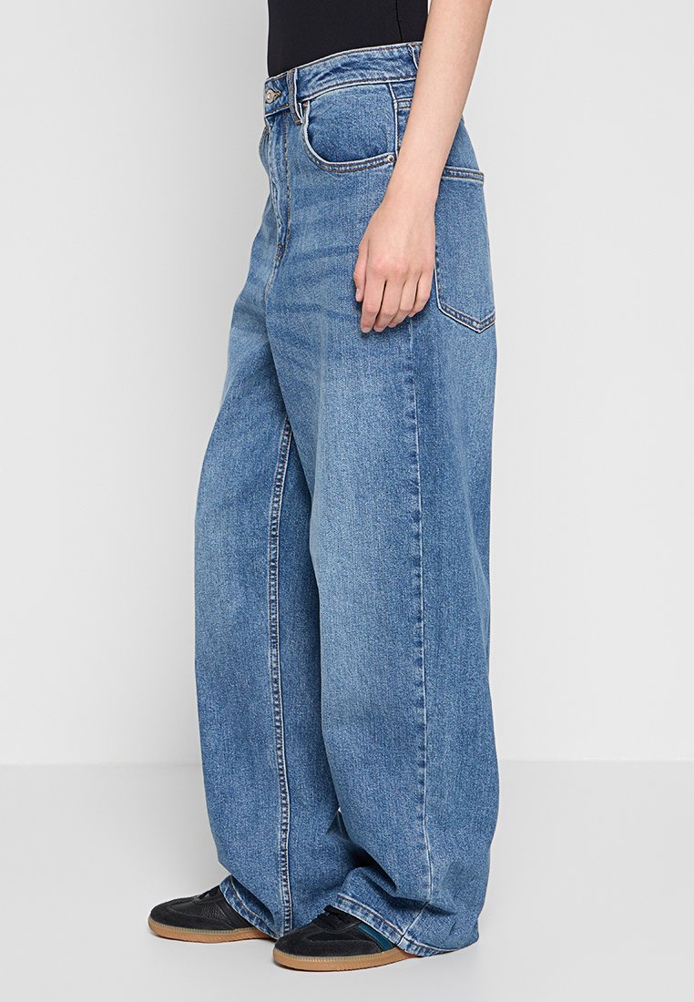 Weite blaue Denim-Jeans mit hoher Taille und Vordertaschen. Der Stoff hat eine ausgebleichte Optik. Kombiniert mit schwarzen Sneakern auf einem neutralen Hintergrund.