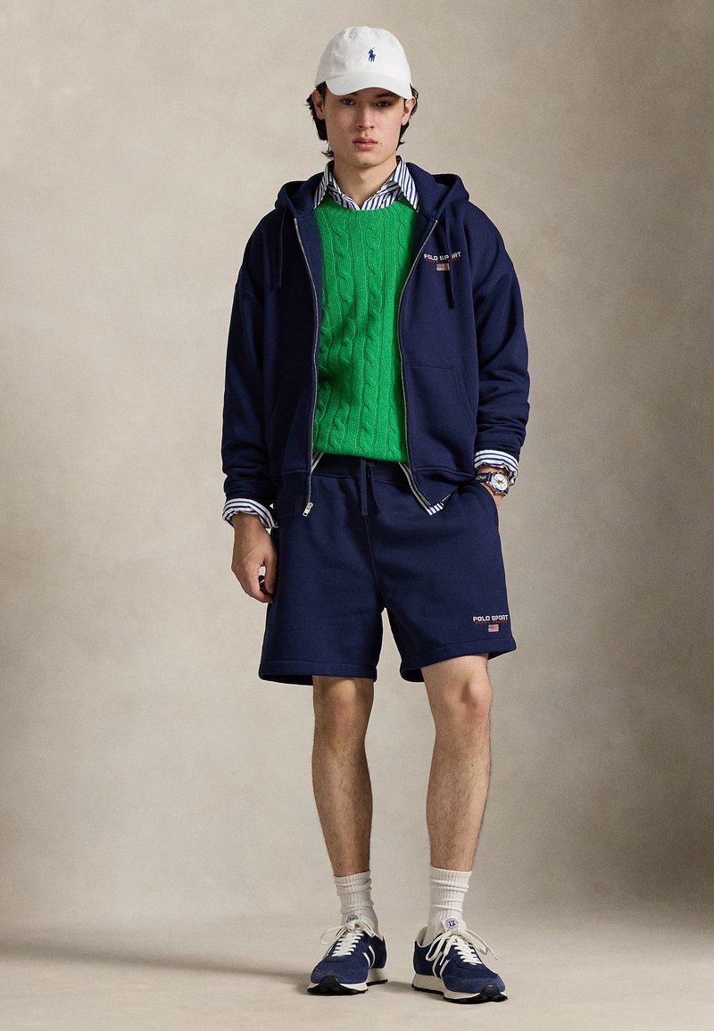 Navy zip-up hoodie, green cable-knit sweater, navy shorts, and white cap. Sneakers feature blue accents. Textured fabrics with stripes visible.