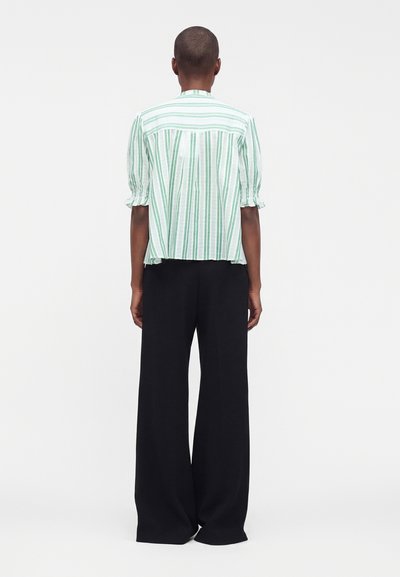 Personne debout, de dos, portant un chemisier à manches courtes rayé vert et blanc et un pantalon noir large et ample.