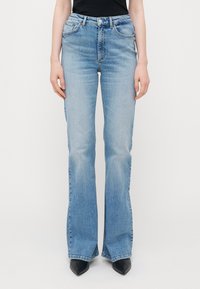 Calças de ganga azuis claras, com corte largo, apresentando cinco bolsos, cintura média e um ligeiro desgaste. A bainha tem um detalhe em fenda.