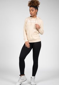 Beige hoodie met "GORILLA WEAR"-print, voorzien van een voorzak en trekkoordjes, gecombineerd met zwarte leggings en witte sportschoenen.