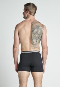 Svarta kalsonger med en grå randig midjeband, bärs av en manlig modell. Anmärkningsbar tatuering av ett lejon på ryggen. Slät textur.