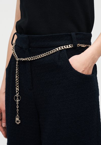 Άτομο που φοράει μαύρο παντελόνι με υφή και ζώνη από χρυσή αλυσίδα με ένα μικρό κλειδαριά-γούρι που κρέμεται μπροστά.