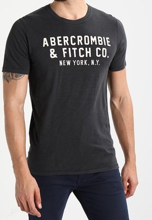 Mężczyzna ubrany w dopasowaną czarną koszulkę z nadrukiem "Abercrombie & Fitch Co. New York, N.Y." z przodu, w połączeniu z ciemnoniebieskimi spodniami.