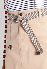 Pantalones beiges con un cinturón trenzado a rayas atado a la cintura, con borlas. La tela es suave con una ligera textura y sutiles detalles de costura.