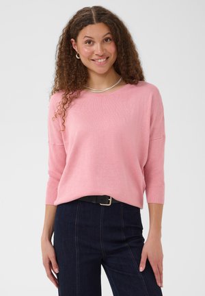Jeune femme aux cheveux bouclés souriante, portant un pull rose à manches trois-quarts et un jean taille haute bleu foncé avec une ceinture noire.