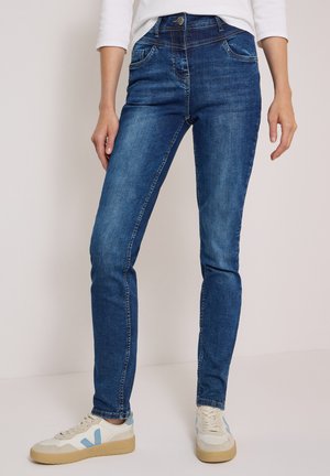 Personne portant un jean skinny bleu, des baskets blanches avec des touches bleu clair et un haut blanc à manches longues, debout devant un fond uni.