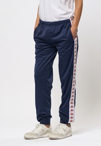 Person som bär marinblå träningsbyxor med vita och röda logostripes på sidorna och vita sneakers, står med händerna i fickorna.