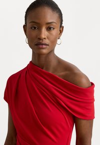 Rochie roșie, cu umerii descoperiți, din material plisat, cu mâneci scurte, textură fină și luciu subtil. Accesorizată cu o cercei micuți din cerc.