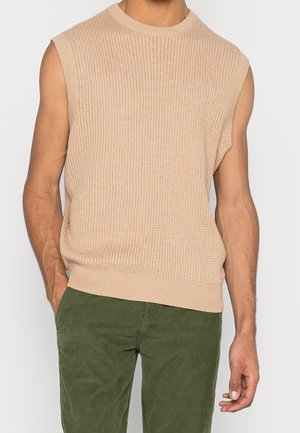 Pull sans manches en maille beige porté par une personne associé à un pantalon en velours côtelé vert sur un fond uni.