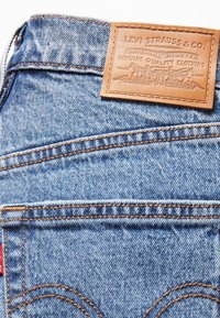 Jean en denim avec couture orange et une étiquette en cuir beige Levi Strauss & Co. au-dessus de la poche arrière sur un tissu bleu clair.