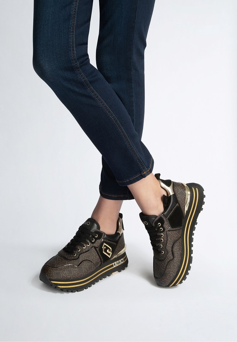 Personne portant un jean skinny bleu foncé et des baskets marron à motifs avec des accents dorés, debout sur un sol gris clair.