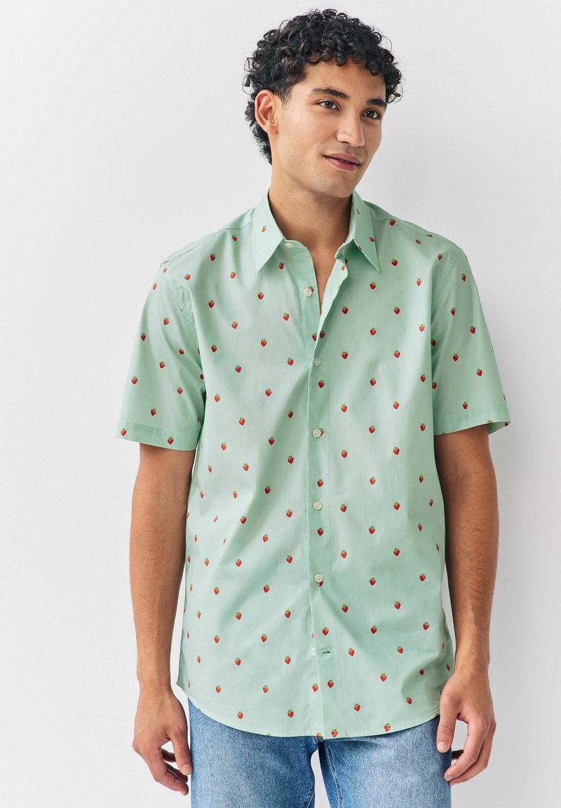 Joven con pelo rizado que lleva una camisa de botones de manga corta de color verde claro con pequeños patrones de fresas rojas y jeans azules.
