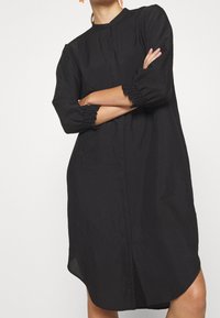 Femme portant une robe noire à manches longues, ample, avec des poignets froncés et un ourlet arrondi, les bras croisés devant un fond blanc.