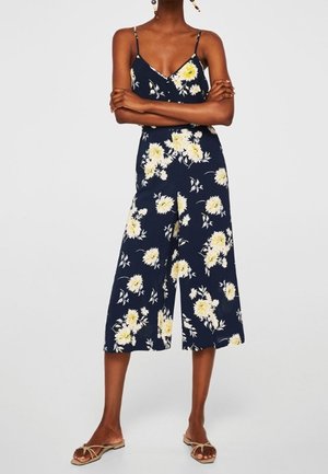 Kvinde iført en mørkeblå ærmeløs buksedragt med gul- og hvidt blomsterprint og beige flade sandaler, stående med armene over kors.