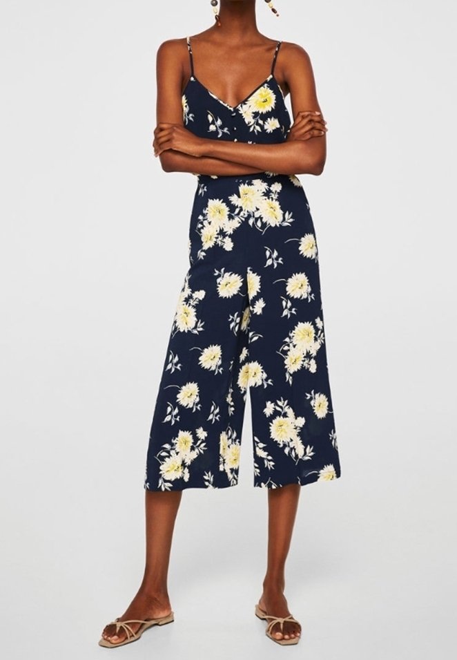 Femme portant une combinaison bleu marine sans manches avec un imprimé floral jaune et blanc et des sandales plates beiges, debout les bras croisés.