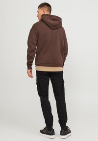 Brun huvtröja med huva, bärs över ett beige lager, i kombination med svarta cargobyxor och svarta sneakers med vita sulor.