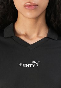 Detailný záber na krk a pery osoby oblečenej v čiernom tričku s výstrihom do V a bielym vyšívaným logom Fenty Puma na hrudi.