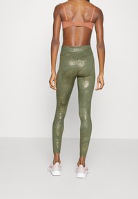 Leggings verdi con motivo glitterato dorato, vita alta e forma aderente, abbinati a un reggiseno sportivo marrone e scarpe da ginnastica rosa chiaro.