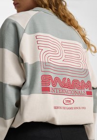 Gestreepte sweatshirt in lichtgroen en crème, met opvallende rode graphics waarop staat "23 SWARM INTERNATIONALS" en "SERVING THE GAME SINCE 1923."